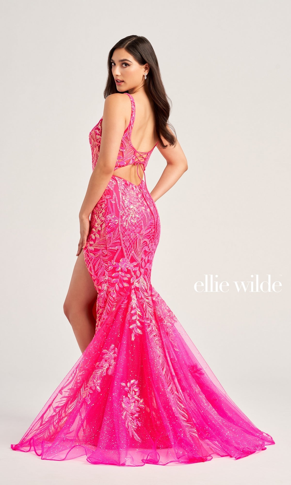 A woman poses in the Sequin Ellie Wilde Sleek Long Prom Dress EW35201—a bright pink mermaid gown with sequins, floral patterns, a lace-up open back, and flared train. The